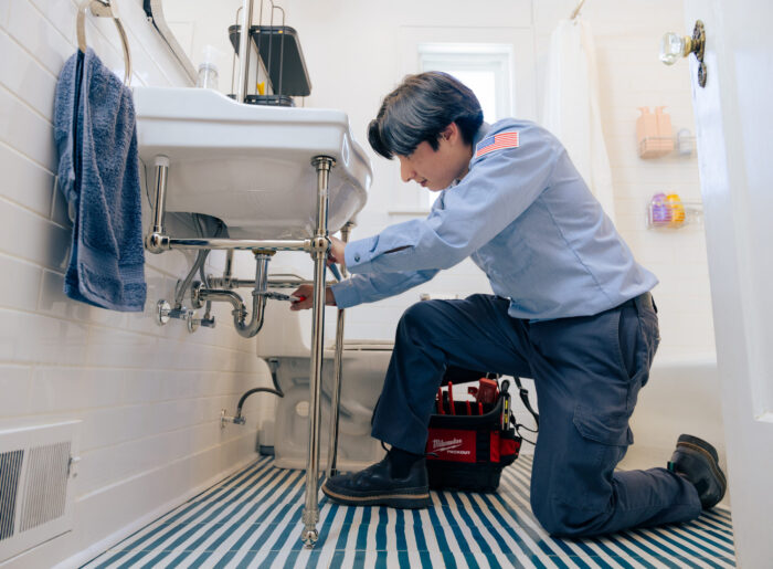 Tacoma plumber kneeling in a bathroom, using pliers to repair the exposed pipes and P-trap under a pedestal sink