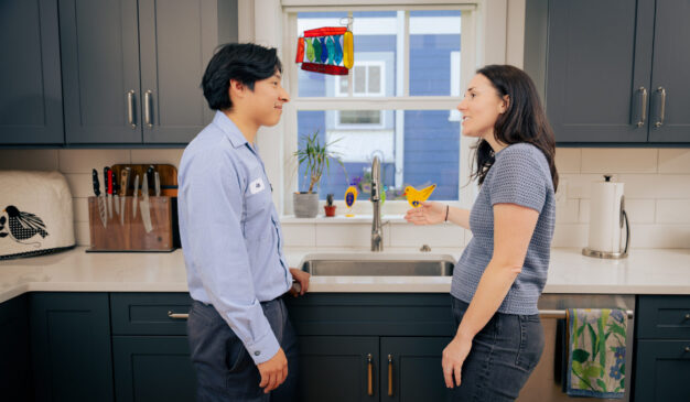 Pacific Heating & Cooling plumber talking with a homeowner at a running kitchen faucet in a Tacoma home